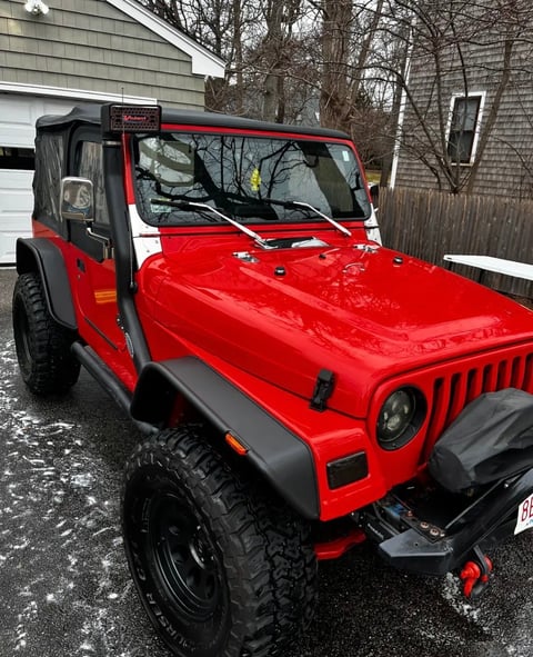 Bright red Jeep Wrangler with black soft top parked in a driveway with bare trees and houses in background