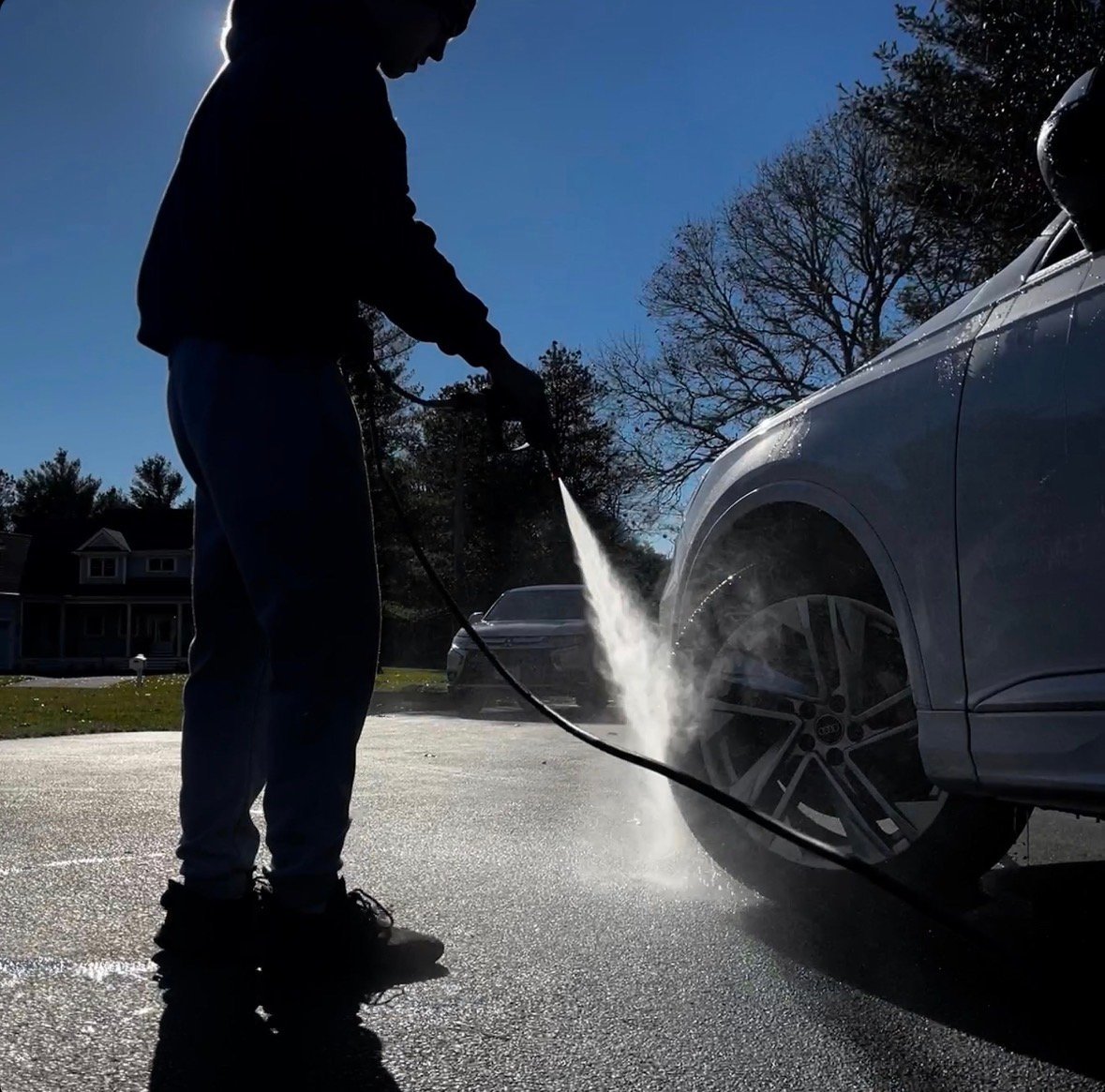 Person washing the side of a silver car with a high-pressure water hose on a residential driveway